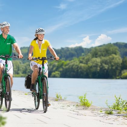 Zwei Radfahrer fahren auf einem gepflasterten Weg entlang der Donau. Grüne Hügel und blauer Himmel im Hintergrund.