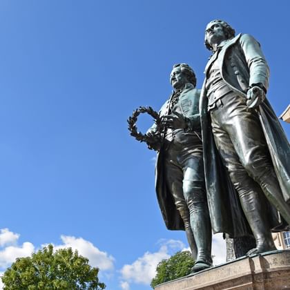 Bronzestatue von Goethe und Schiller, die zusammen auf einem Sockel vor einem klassischen Gebäude unter blauem Himmel mit weißen Wolken stehen.