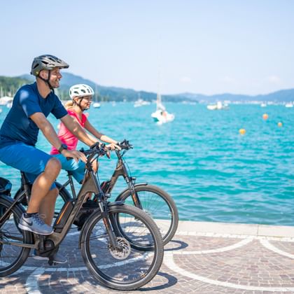 Zwei Radfahrer mit Helmen fahren E-Bikes entlang einer Seepromenade. Türkisfarbenes Wasser mit Segelbooten und grünen Hügeln im Hintergrund.
