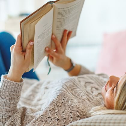 Frau in beigem Pullover und blauen Socken liegt auf dem Bett und liest ein aufgeschlagenes Buch. Rosa Kissen im Hintergrund sichtbar.
