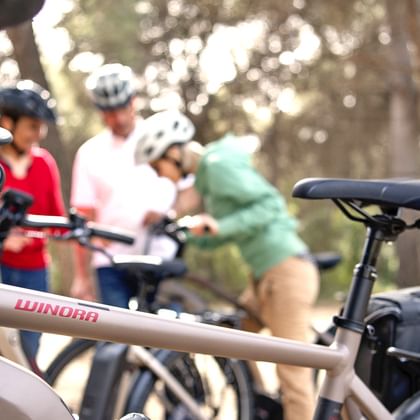 Nahaufnahme eines weißen E-Bikes mit Winora-Marke im Vordergrund, mit unscharfer Radfahrergruppe mit Helmen im Hintergrund zwischen Bäumen.