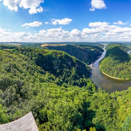 Panoramablick auf die Saarschleife durch dicht bewaldete Hügel. Aussichtsplattform links sichtbar unter blauem Himmel.