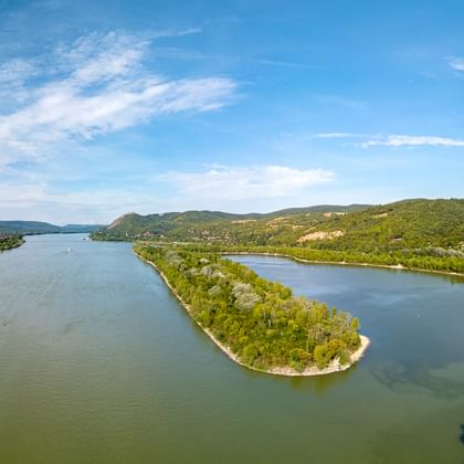 Danube River bend with forested island Aerial view of the Danube River forming a wide bend around a forested island, surrounded by green hills under a blue sky with white clouds.