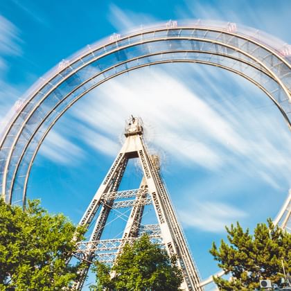 Langzeitbelichtung des Wiener Prater Riesenrads in Bewegung, mit kreisförmiger Unschärfe vor blauem Himmel, umgeben von grünen Bäumen.