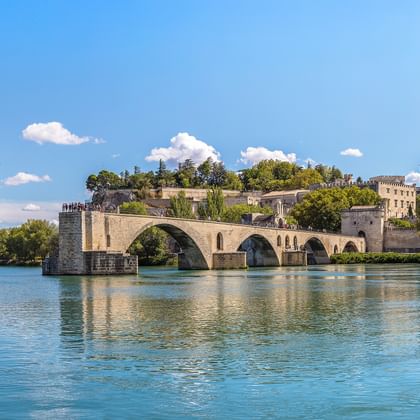 Historische Steinbrücke mit Bögen über die Rhône, mit mittelalterlichem Palast und Turm am gegenüberliegenden Ufer unter blauem Himmel.