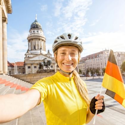 Lächelnde Radfahrerin in gelbem Trikot und Helm mit deutscher Flagge am Gendarmenmarkt in Berlin mit Französischem Dom im Hintergrund.