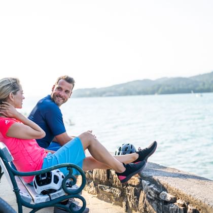 Zwei Radfahrer sitzen auf einer Steinmauer an einem Kärntner See, mit Fahrrädern neben sich und bewaldeten Bergen im Hintergrund.