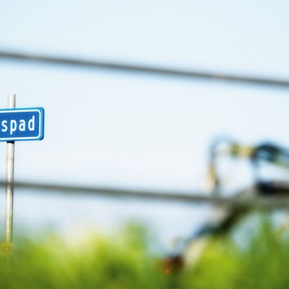 Blaues Fahrradweg-Schild mit der Aufschrift 'fietspad' und einem unscharfen Radfahrer im Hintergrund an einem sonnigen Tag in Friesland.