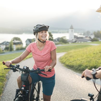 Zwei Radfahrer mit Helmen auf einem asphaltierten Weg in Oberösterreich. Grüne Wiesen und ein Seeufer-Dorf mit Kirchturm im Hintergrund.