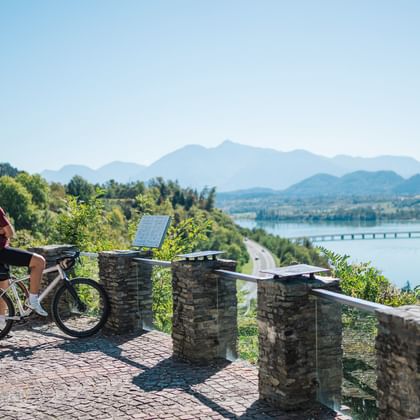 Radfahrer mit E-Bike an steinerner Aussichtsplattform mit Blick auf Fluss, Brücke und Berge. Klarer blauer Himmel und grüne bewaldete Hügel.