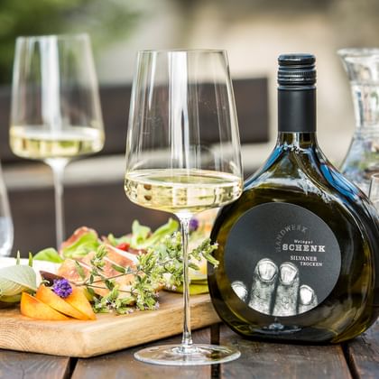 Tisch im Freien mit Weißweingläsern, dunkler Weinflasche, Wasserkaraffe und Holzbrett mit Käse und essbaren Blüten auf Holzlatten.