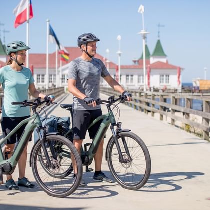 Two cyclists with e-bikes standing on a wooden boardwalk leading to a historic pier building with flags, blue sea and sky in background.