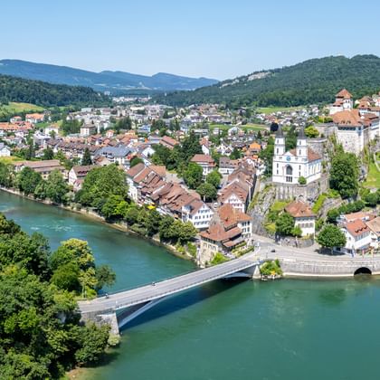 Luftaufnahme einer Schweizer Stadt entlang der türkisfarbenen Aare mit Steinbrücke, traditionellen Gebäuden, Kirchturm und bewaldeten Hügeln.