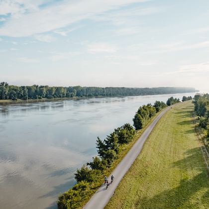 Luftaufnahme von zwei Radfahrern auf einem asphaltierten Weg entlang eines breiten Flusses. Grüner Damm trennt den Weg vom Wasser, Wälder auf beiden Seiten.