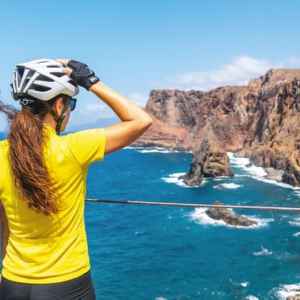 Radfahrerin in gelbem Trikot und weißem Helm betrachtet Madeiras felsige Küste mit türkisfarbenem Ozean und dramatischen Klippen.