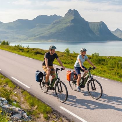 Zwei Radfahrer mit Packtaschen auf einer Küstenstraße in Norwegen mit dramatischen Berggipfeln und Fjord im Hintergrund unter blauem Himmel.