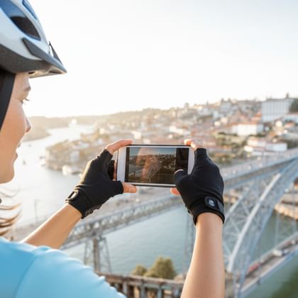Radfahrerin mit Helm fotografiert mit Smartphone historische Brücke über den Douro mit Porto-Stadtbild im Hintergrund.