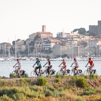 Gruppe von Radfahrern auf einem Küstenweg mit mediterranem Hafen und historischer Hügelstadt im Hintergrund. Segelboote liegen in der Bucht.