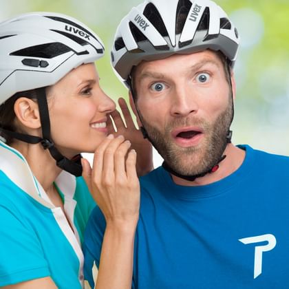 Frau in türkisfarbenem Shirt und weißem Helm flüstert einem bärtigen Mann in blauem Shirt und weißem Helm zu, der überrascht aussieht.