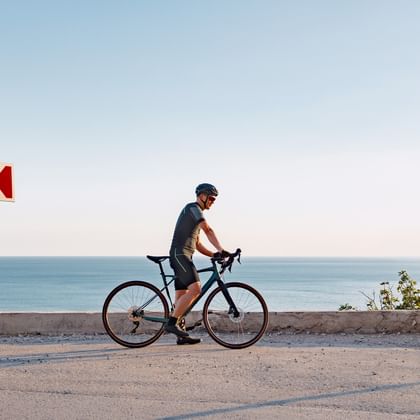 Radfahrer mit Rennrad auf Küstenstraße in der Algarve mit Meerblick, rot-weißem Verkehrsschild und klarem blauen Himmel.