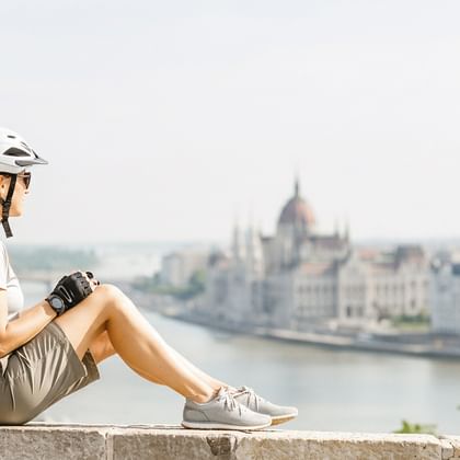 Radfahrerin mit Helm sitzt auf Steinmauer mit Blick auf die Donau und das ungarische Parlamentsgebäude in Budapest.