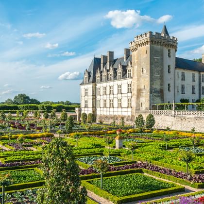 Weißes Steinschloss mit Turm und formalen Gärten mit geometrischen Hecken, bunten Blumenbeeten und gepflegten Rasenflächen unter blauem Himmel.