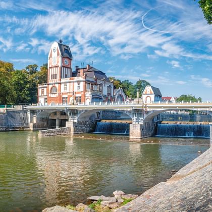 Steinbrücke mit Wehr über die Elbe, mit rosa Gebäude und Uhrturm. Bäume rahmen die Szene unter blauem Himmel ein.