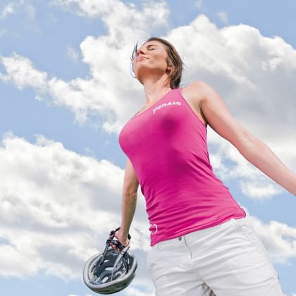Frau in pinkem Radtrikot und weißer Shorts hält einen Helm, steht vor blauem Himmel mit weißen Wolken, Augen geschlossen, genießt den Moment.