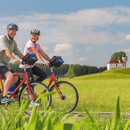 Zwei Radfahrer mit Packtaschen fahren durch grüne Felder in Bodensee-Oberschwaben. Eine kleine Kapelle auf einem Hügel und Wald im Hintergrund unter blauem Himmel.