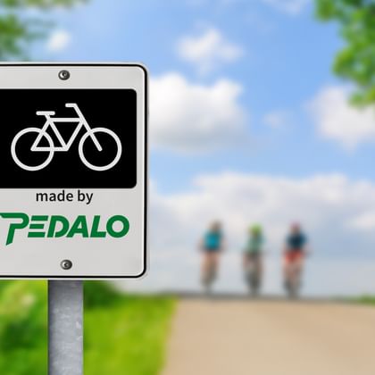 White sign with bicycle symbol and 'made by PEDALO' text on a cycling path. Three cyclists visible in the blurred background under blue sky.