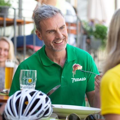 Mann im grünen Pedalo-Poloshirt mit Tablet spricht mit Radfahrerin im gelben Shirt im Straßencafé. Fahrradhelm auf dem Tisch sichtbar.