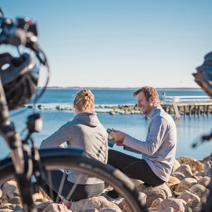 Zwei Radfahrer mit Rädern rasten auf felsigem Ufer am ruhigen blauen Wasser an Schwedens Westküste. Alte Holzpfähle im Hintergrund.
