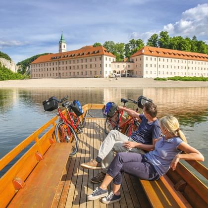 Two cyclists with bikes on wooden ferry crossing the Danube River. Historic monastery with orange roof and green tower visible on riverbank.