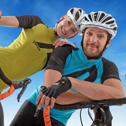 Two smiling cyclists wearing helmets and cycling gear pose with their bikes against a blue sky. One wears a yellow jersey, the other turquoise.