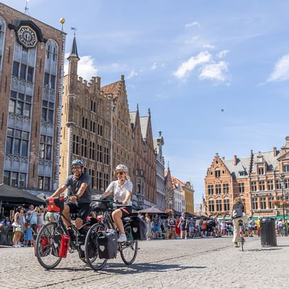 Zwei Radfahrer fahren über einen Kopfsteinpflasterplatz in Brügge, umgeben von historischen flämischen Gebäuden mit Treppengiebeln und Glockenturm.