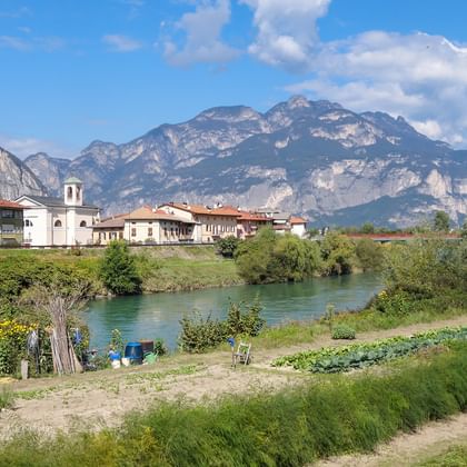 Dorf mit Kirchturm neben türkisfarbener Etsch, umgeben von dramatischen Alpenbergen unter blauem Himmel mit weißen Wolken.