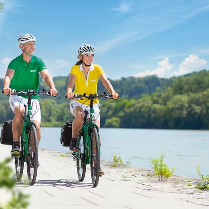 Zwei Radfahrer mit Helmen auf grünen Rädern auf einem Weg am See. Einer trägt ein grünes Shirt, der andere gelb. Hügel und Bäume im Hintergrund.