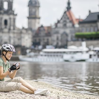 Radfahrerin mit Helm sitzt am Flussufer mit gelbem Rucksack und hält eine Kamera. Dresdner Altstadtpanorama mit Kirchen an der Elbe.