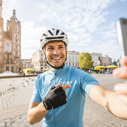 Lächelnder Radfahrer in blauem Trikot und weißem Helm macht Selfie mit Smartphone auf historischem Stadtplatz mit gotischen Kirchtürmen im Hintergrund.