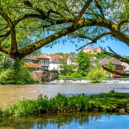 View of the Lahn River framed by large tree branches, with traditional half-timbered houses and a castle on a hill in the background.