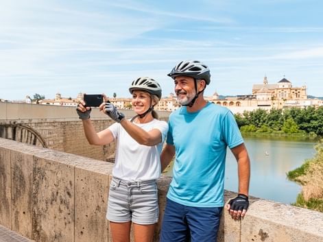Zwei Radfahrer mit Helmen machen ein Selfie auf einer Steinbrücke in Andalusien. Historische Gebäude und ein Fluss sind im Hintergrund sichtbar.