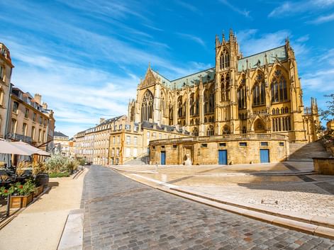 Gotische Kathedrale mit verzierten Türmen in Metz, Frankreich. Das gelbe Steingebäude überblickt einen gepflasterten Platz mit Wohngebäuden links.