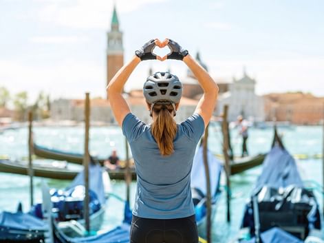 Radfahrerin mit Helm formt Herz mit Händen vor Venedigs Lagune mit Gondeln und dem Campanile von San Giorgio Maggiore.