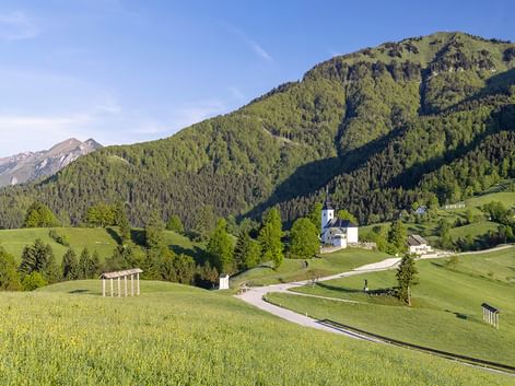 Weiße Kirche mit Glockenturm in grünem Tal umgeben von sanften Wiesen und bewaldeten Bergen unter blauem Himmel in Slowenien.