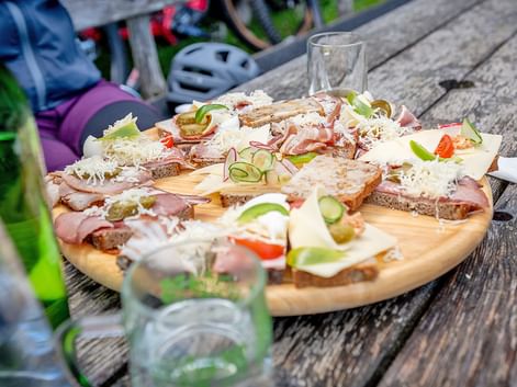 Holzbrett mit belegten Broten mit Schinken, Käse, Gurke und Gemüse auf Picknicktisch. Fahrradausrüstung und Helme sichtbar.