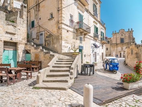 Kopfsteinpflasterplatz in Apulien mit Steintreppe, verwitterten Gebäuden, blauer Ape und barocker Kirche unter blauem Himmel.