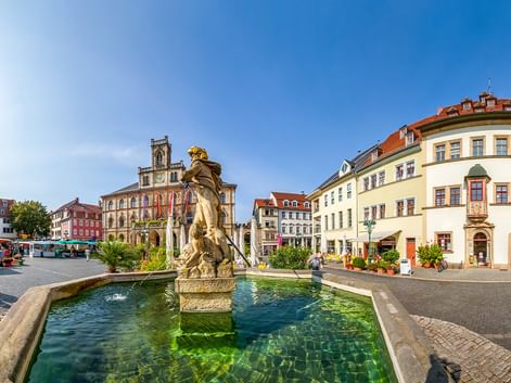 Prunkvoller Barockbrunnen mit goldener Statue in der Mitte eines Kopfsteinpflaster-Marktplatzes umgeben von bunten historischen Gebäuden.