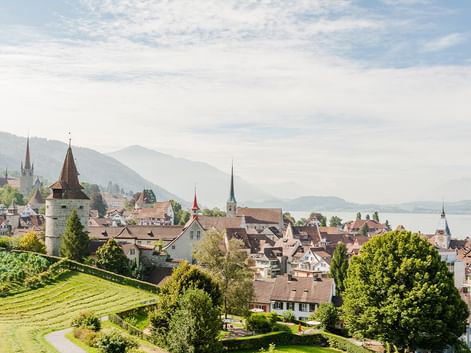 Schweizer Stadt am Vierwaldstättersee mit historischen Kirchtürmen und traditionellen Gebäuden, umgeben von grünen Hügeln und Bergen.