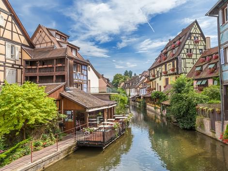 Schmaler Kanal zwischen bunten Fachwerkhäusern im Süd-Elsass. Holzterrasse ragt über das Wasser, üppiges Grün säumt die Ufer.
