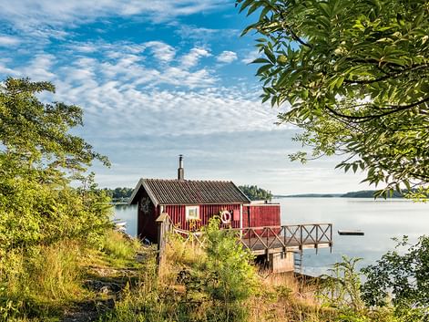 Rotes Holz-Bootshaus mit Wellblechdach und Holzsteg, der sich in ruhiges Seewasser erstreckt, umgeben von grünen Bäumen unter bewölktem Himmel.